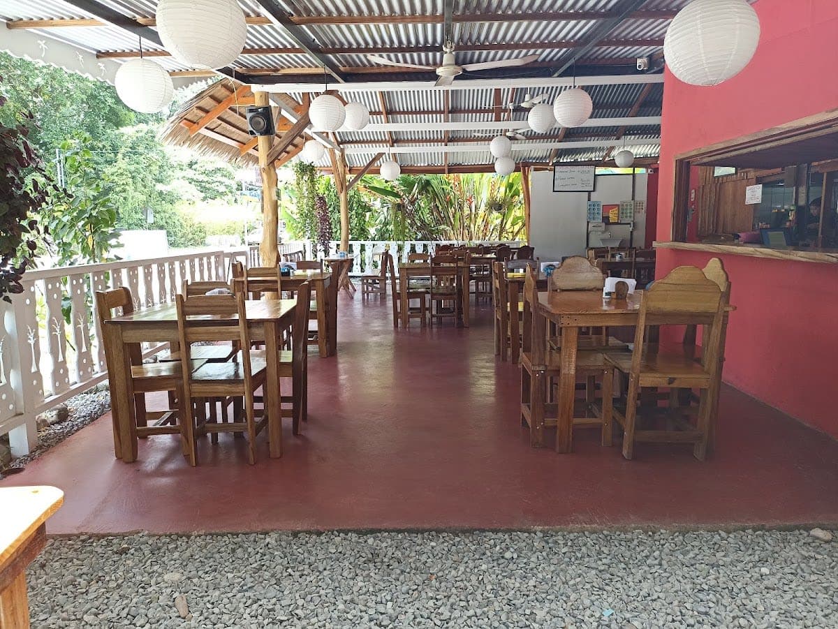 A weathered wooden table holds an overflowing partitioned plate of golden turmeric-spiced coconut rice and beans alongside dark, glossy stewed Caribbean chicken, with a small cup of vibrant passionfruit sauce catching the humid afternoon light filtering through an open wall overlooking the soft-focus jungle beyond.